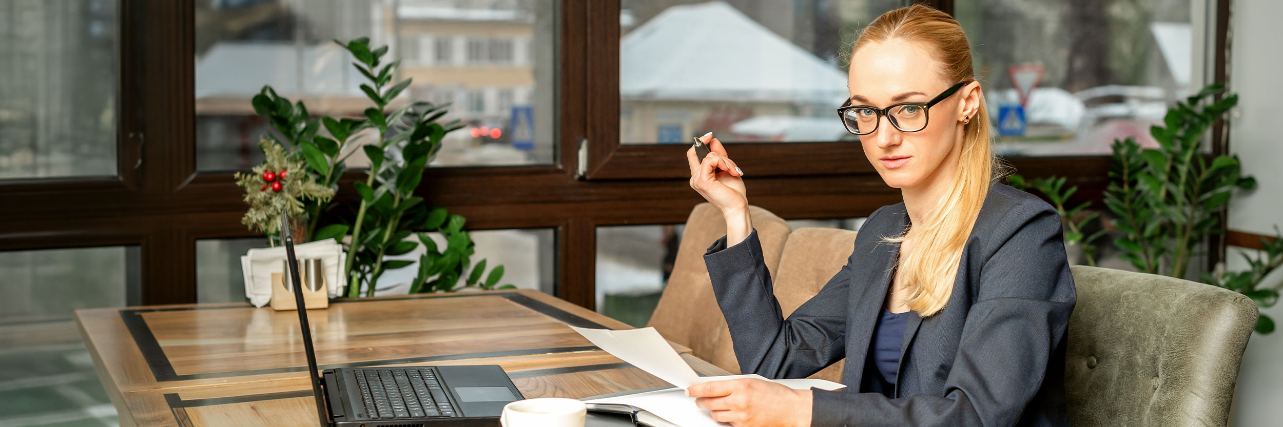 FOA_desktop Front office administrator wearing glasses and a blazer reviewing documents at a desk with a laptop in a professional office setting.