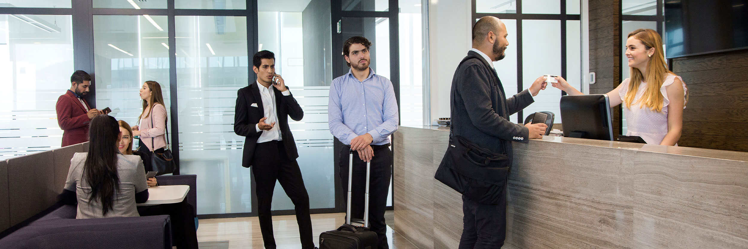 HM-CooP Hotel receptionist assisting a guest at the front desk while other business travelers wait and interact in a modern hotel lobby.