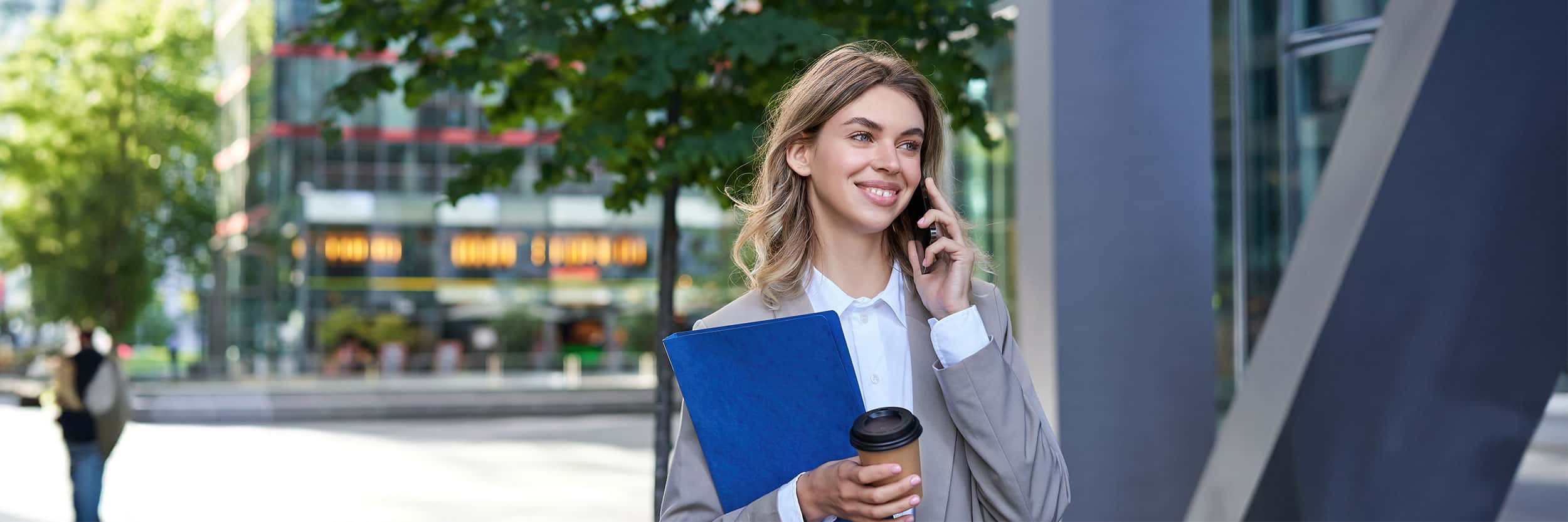 IA Smiling business professional in a grey blazer talking on her phone while holding a blue folder and coffee outside a modern office building.