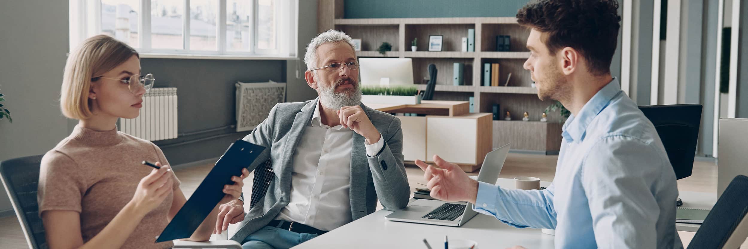 IP Three business professionals discussing documents during a meeting in a modern office, with a laptop and clipboard on the table.