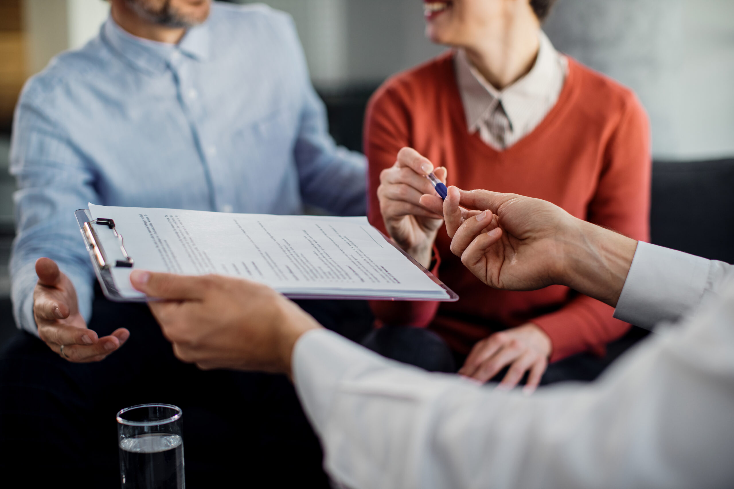 Close-up of hands exchanging a pen over a clipboard with documents, with two people sitting across a desk in a meeting.