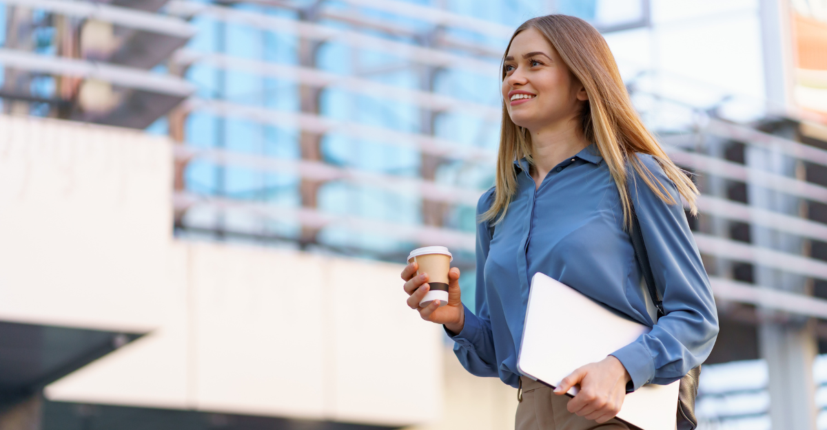 A young woman with long blonde hair, dressed in a blue blouse and beige pants, walking outside near a modern glass building. She is smiling, holding a coffee cup in one hand and a laptop in the other.