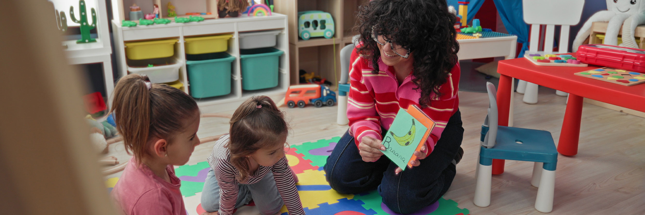 ECEA_desktop Early childhood educator reading a book to two young children during a play-based learning activity in a colourful classroom.