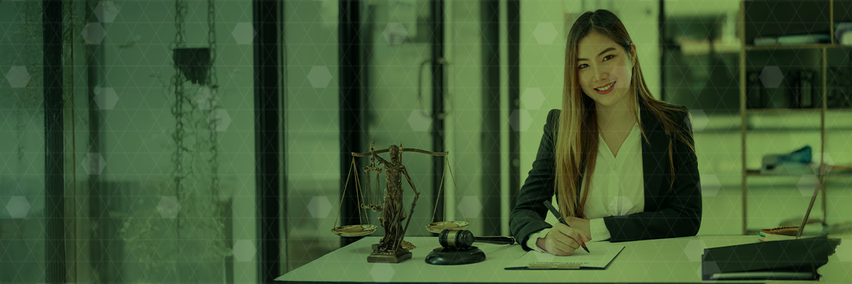 paralegal-banner-1 Smiling female lawyer sitting at a desk in a modern office, with a scales of justice statue and gavel beside her.
