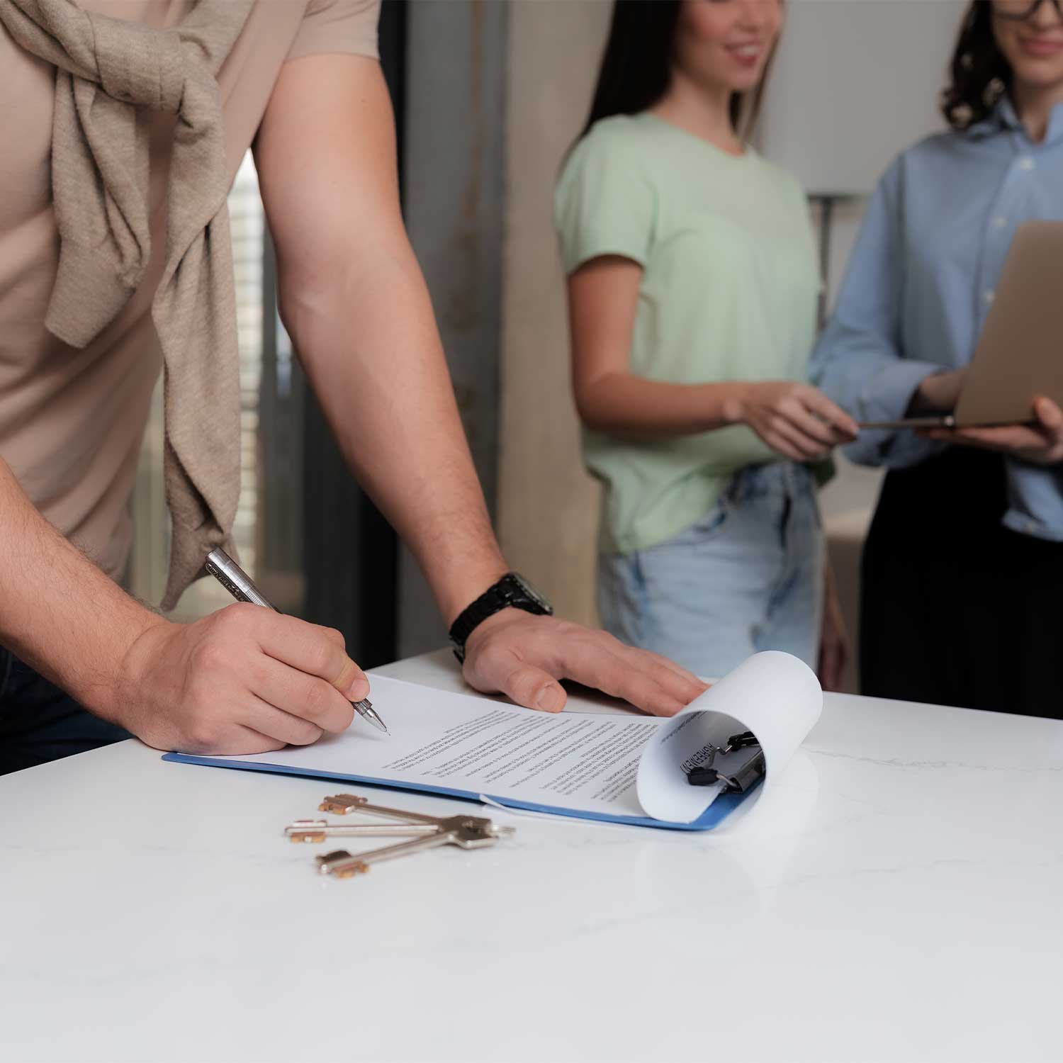 Person signing a contract with keys on the table while others review documents in the background
